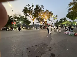 Bức ảnh chụp cảnh phố đi bộ vào buổi tối, có lẽ là khu vực quanh Hồ Gươm ở Hà Nội. Bầu trời đã về chiều, đèn đường và đèn trang trí trên các cây cổ thụ đã bật sáng, tạo nên một không gian ấm cúng. Rất đông người dân và du khách đang tản bộ trên con đường rộng đã được chặn xe. Mọi người mặc trang phục đa dạng, có các gia đình, cặp đôi và nhóm bạn trẻ. Ở phía xa, có thể thấy bóng dáng của một công trình kiến trúc cổ, có cờ đỏ sao vàng đang bay. Bên phải đường, một người bán hàng rong đang bày bán rất nhiều thú nhồi bông đủ màu sắc. Một phần ngón tay của người chụp ảnh vô tình lọt vào khung hình ở góc trên bên trái.。（日本語訳）。夜の歩行者天国の風景を撮影した写真で、おそらくハノイのホアンキエム湖周辺のエリアでしょう。空は夕暮れ時になり、街灯や古い木々に飾られたイルミネーションが灯り、温かみのある空間を創り出しています。車両通行止めになった広い道を、多くの地元の人々や観光客が散策しています。人々は様々な服装をしており、家族連れ、カップル、そして若い友人たちのグループがいます。遠くには、古い建築物のシルエットが見え、金星紅旗がはためいています。道の右側では、露天商が色とりどりのぬいぐるみをたくさん並べて売っています。撮影者の指の一部が、誤って左上の角に写り込んでいます。