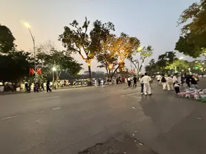Bức ảnh chụp cảnh một con đường đi bộ đông đúc vào buổi chiều tối, có lẽ là khu vực quanh Hồ Gươm ở Hà Nội. Bầu trời đã nhá nhem tối và đèn đường đã được bật sáng, tạo ra ánh sáng vàng ấm áp hắt lên những tán cây cổ thụ ven đường. Rất nhiều người, bao gồm các gia đình, cặp đôi và nhóm bạn, đang thong thả đi dạo trên con đường rộng. Ở phía xa, có thể nhìn thấy một công trình kiến trúc cổ và những lá cờ đỏ. Bên phải đường, có một người bán hàng rong đang bày bán những con thú nhồi bông màu hồng và trắng.。（日本語訳）。写真は夕方の混雑した歩道の光景を写したもので、おそらくハノイのホアンキエム湖周辺のエリアでしょう。空はすでに薄暗く、街灯が灯され、道端の古い木々の葉に暖かい黄色の光を投げかけています。家族、カップル、友人グループを含む多くの人々が、広い道をのんびりと散歩しています。遠くには、古い建築物と赤い旗が見えます。道の右側には、ピンクと白のぬいぐるみを並べて売っている露天商がいます。