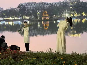 Một cặp đôi đang chụp ảnh bên bờ Hồ Gươm vào buổi chiều tà. Chàng trai mặc áo dài nam màu trắng và quần đen, trên vai cầm một bó cành lá xanh, mỉm cười nhìn về phía máy ảnh. Cô gái mặc áo dài trắng, quay lưng lại và cúi người tạo dáng, mái tóc đen dài được buộc cao. Phía sau họ là khung cảnh Hồ Gươm thơ mộng với Tháp Rùa cổ kính được chiếu đèn vàng rực rỡ, in bóng xuống mặt nước. Xa xa là những tòa nhà và hàng cây ven hồ cũng đã lên đèn. Bầu trời có màu hồng nhạt của hoàng hôn.。（日本語訳）。あるカップルが夕暮れ時のホアンキエム湖のほとりで写真を撮っている。男性は白い男性用アオザイと黒いズボンを着用し、肩に緑の葉の枝束を担ぎ、カメラに向かって微笑んでいる。女性は白いアオザイを着て、後ろを向き、身をかがめてポーズをとっており、長い黒髪は高く結われている。彼らの後ろには、鮮やかな黄色のライトで照らされ、水面に影を落とす古風な亀の塔がある、詩的なホアンキエム湖の風景が広がっている。遠くには、建物や湖畔の並木も見え、それらもライトアップされている。空は夕焼けの薄ピンク色をしている。