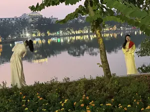 Khung cảnh buổi tối bên hồ, có hai người phụ nữ mặc áo dài truyền thống. Người phụ nữ bên trái mặc áo dài màu kem, đang tạo dáng cúi người xuống. Người phụ nữ bên phải mặc áo dài màu vàng, quàng khăn đỏ, tay cầm một chiếc quạt. Phía sau họ là mặt hồ rộng, phản chiếu ánh đèn từ thành phố bên kia bờ. Ở tiền cảnh có một luống hoa màu vàng và cam.。（日本語訳）。湖畔の夜景、伝統的なアオザイを着た二人の女性がいます。左の女性はクリーム色のアオザイを着て、身をかがめるポーズをしています。右の女性は黄色のアオザイを着て、赤いスカーフを巻き、手に扇子を持っています。彼女たちの後ろは広い湖面で、対岸の街からの灯りを反射しています。前景には黄色とオレンジ色の花壇があります。