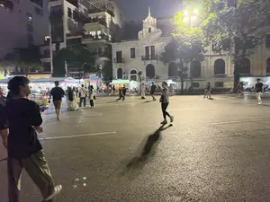 Bức ảnh chụp cảnh một quảng trường hoặc một con phố rộng ở thành phố vào ban đêm. Mặt đường nhựa ẩm ướt, phản chiếu ánh sáng vàng từ những ngọn đèn đường. Nhiều người đang đi dạo, tản bộ trong không gian thoáng đãng. Ở phía trước, một chàng trai trẻ mặc áo phông đen và quần rộng. Ở giữa, một người phụ nữ đang vừa đi vừa nhìn vào điện thoại. Phía sau là những tòa nhà với nhiều phong cách kiến trúc khác nhau, nổi bật là một tòa nhà cổ màu trắng kem. Xa hơn là các cửa hàng, gian hàng sáng đèn, tạo nên một khung cảnh nhộn nhịp về đêm.。（日本語訳）。写真は、夜の街の広場か広い通りの風景を写しています。アスファルトの路面は湿っており、街灯の黄色い光を反射しています。広々とした空間で、多くの人々が散歩したり、ぶらぶら歩いたりしています。手前には、黒いTシャツとゆったりしたズボンを履いた若い男性がいます。中央では、一人の女性が歩きながらスマートフォンを見ています。後ろには様々な建築様式の建物があり、特にクリーム色の古い建物が目立っています。さらに奥には明かりが灯った店や屋台があり、夜の賑やかな光景を作り出しています。