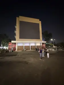 Cảnh quảng trường vào ban đêm. Ở trung tâm là một tòa nhà lớn với một màn hình điện tử khổng lồ đang tắt. Nhiều người, bao gồm cả người lớn và trẻ em, đang đi dạo và tụ tập trong quảng trường. Một bé gái mặc bộ đồ màu sáng đang đứng ở phía trước. Bên trái tòa nhà có một tấm pano lớn màu đỏ với hình ngôi sao vàng, giống quốc kỳ Việt Nam. Không gian được chiếu sáng bởi đèn đường và có cây cối xung quanh.。（日本語訳）。夜の広場の光景。中央には、電源が切れている巨大な電光掲示板がある大きな建物があります。大人や子供を含む多くの人々が、広場を散策したり集まったりしています。明るい色の服を着た一人の女の子が手前に立っています。建物の左側には、ベトナム国旗のような、黄色い星の描かれた大きな赤いパネルがあります。空間は街灯で照らされており、周りには木々があります。