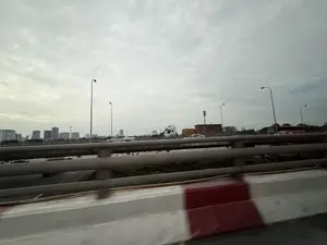 Khung cảnh được chụp từ một phương tiện đang di chuyển trên cầu vượt hoặc đường cao tốc. Bầu trời u ám, bao phủ bởi những đám mây xám. Trên cầu có nhiều loại xe cộ đang lưu thông, bao gồm ô tô con và xe tải. Phía dưới cầu vượt là một con đường khác cũng có xe đang chạy. Ở phía xa là quang cảnh thành phố với nhiều tòa nhà cao tầng. Dọc theo đường là các cột đèn chiếu sáng. Ở tiền cảnh, dải phân cách bê tông bị mờ do chuyển động.。（日本語訳）。光景は、高架橋または高速道路を移動中の乗り物から撮影されたものです。空は曇っており、灰色の雲に覆われています。橋の上では、乗用車やトラックを含む多くの種類の車両が通行しています。高架橋の下には、同じく車が走っている別の道路があります。遠くには、多くの高層ビルがある街の景色が見えます。道路に沿って街灯があります。前景では、コンクリートの中央分離帯が動きによってぼやけています。