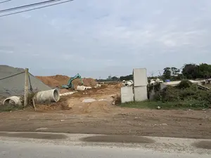 Đây là hình ảnh một công trường xây dựng. Ở giữa là một chiếc máy xúc màu xanh lá cây đang làm việc trên nền đất bùn. Xung quanh có nhiều đống đất, sỏi đá và các ống cống bê tông lớn nằm rải rác. Bên phải có các khối bê tông hình chữ nhật được xếp chồng lên nhau. Phía xa là cây cối và các tòa nhà đang được xây dựng với cần cẩu. Bầu trời u ám và có nhiều mây.。（日本語訳）。これは建設現場の写真です。中央には、緑色のショベルカーが泥の上で作業しています。周りには、土や砂利の山、そして大きなコンクリート製の土管が散らばっています。右側には、長方形のコンクリートブロックが積み重ねられています。遠くには、木々や、クレーンで建設中の建物があります。空はどんよりとしていて、雲が多いです。