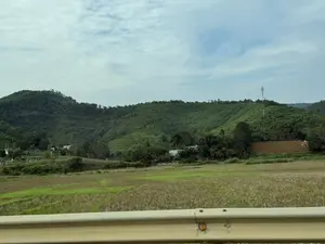 Bức ảnh chụp phong cảnh một vùng nông thôn miền núi. Phía sau là những ngọn đồi trập trùng được bao phủ bởi cây cối xanh tươi, rậm rạp. Dưới chân đồi, có một vài ngôi nhà nhỏ nằm xen kẽ giữa các lùm cây. Trên một đỉnh đồi, có một cột thu phát sóng viễn thông. Phía trước là một cánh đồng rộng, một phần đã được thu hoạch để lại gốc rạ màu nâu vàng, phần còn lại vẫn còn màu xanh của cây cỏ. Bầu trời có nhiều mây trắng. Ở gần nhất là một phần của lan can bảo vệ đường bộ màu vàng.。（日本語訳）。山岳地帯の田舎の風景を写した写真です。後方には、青々とした木々が生い茂る、起伏のある丘が連なっています。丘の麓には、木々の間にいくつかの小さな家が点在しています。丘の頂上の一つには、通信用の送受信アンテナ塔があります。前方には広い田畑が広がり、一部は収穫されて黄褐色の切り株が残り、残りの部分はまだ草木の緑色をしています。空には白い雲が多く浮かんでいます。最も手前には、黄色い道路のガードレールの一部が見えます。