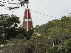 Bức ảnh chụp một tháp chuông nhà thờ có kiến trúc hiện đại, nhô lên từ phía sau những tán cây xanh rậm rạp. Phần trên của tháp có mái màu đỏ và đỉnh là một cây thánh giá. Phần thân tháp được xây bằng gạch màu nâu nhạt. Xung quanh là rất nhiều cây cối um tùm. Bầu trời có màu xám và u ám, với vài đường dây điện vắt ngang qua khung cảnh.。（日本語訳）。写真には現代的な建築様式の教会の鐘楼が写っており、緑豊かな木々の梢の後ろから突き出ている。塔の上部は赤い屋根で、頂上には十字架がある。塔の本体は薄茶色のレンガで建てられている。空は灰色で曇っており、何本かの電線が景色を横切っている。