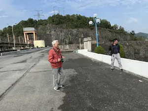 Hai người đàn ông đang đứng trên một con đường nhựa trên đỉnh một con đập. Người đàn ông lớn tuổi bên trái có tóc bạc, đeo kính râm, mặc áo khoác màu đỏ, quần jean màu xám và đang cầm một chiếc máy ảnh. Người đàn ông còn lại ở bên phải đang đứng tựa vào lan can màu trắng, mặc áo khoác phao màu đen và quần màu xám. Phía sau họ là một sườn đồi đá dốc với cây cối xanh tươi và các cột điện cao thế. Bầu trời trong xanh và có mây.。（日本語訳）。二人の男性がダムの頂上にあるアスファルトの道路の上に立っています。左側の年配の男性は白髪で、サングラスをかけ、赤いジャケットとグレーのジーンズを着用し、カメラを手に持っています。右側のもう一人の男性は、白い手すりに寄りかかって立っており、黒いダウンジャケットとグレーのズボンを着用しています。彼らの後ろには、緑豊かな木々と高圧電線の鉄塔がある、急な岩の丘の斜面があります。空は青く澄んでおり、雲があります。
