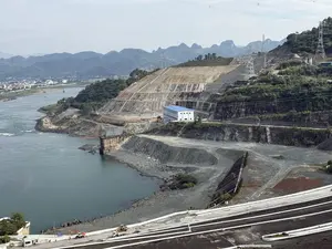 Bức ảnh chụp toàn cảnh một công trình xây dựng quy mô lớn, có thể là một đập thủy điện, nằm bên bờ một con sông rộng. Một sườn núi lớn đã được san ủi và gia cố thành nhiều bậc thang bằng bê tông. Nhiều cột điện cao thế và đường dây điện được dựng lên trên sườn núi. Gần bờ sông, có một tòa nhà màu trắng với mái xanh, có lẽ là nhà máy hoặc trạm điều khiển. Dòng sông có màu nước xanh đậm. Ở phía xa bên kia sông là một thị trấn với nhiều tòa nhà. Phông nền là những dãy núi trập trùng ẩn hiện trong làn sương mờ. Rải rác trong khu vực công trường là những con đường đất, đống đá sỏi và một vài chiếc xe công trình như xe tải và máy xúc.。（日本語訳）。大規模な建設現場の全景写真。おそらく水力発電ダムで、広い川のほとりに位置している。大きな山腹が整地され、コンクリートで複数の段に補強されている。山腹には多くの高圧送電鉄塔と送電線が建てられている。川岸の近くには、青い屋根の白い建物があり、おそらく工場か制御ステーションだろう。川の水は濃い青色をしている。川の向こうの遠くには、多くの建物が建ち並ぶ町がある。背景は、薄い霧の中に現れ隠れする、起伏に富んだ山脈である。建設現場の区域には、未舗装の道路、砂利の山、そしてトラックやショベルカーといった数台の建設車両が点在している。