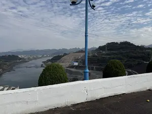 Bức ảnh chụp một khung cảnh rộng lớn từ trên cao, nhìn ra một con sông và vùng núi. Ở phía trước, có một bức tường bê tông màu trắng và một cây cột đèn đường màu xanh lam. Phía xa là một con sông rộng, bên cạnh là một công trình đất lớn có vẻ như là một phần của đập thủy điện, với nhiều đường dây điện cao thế chằng chịt phía trên. Bên kia sông là một thị trấn nhỏ với nhiều tòa nhà. Phông nền là những dãy núi trập trùng dưới bầu trời nhiều mây.。（日本語訳）。写真は、上空から川と山岳地帯を望む広大な風景を写しています。手前には、白いコンクリートの壁と青い街灯柱があります。遠くには広い川があり、その隣には水力発電ダムの一部と思われる大きな土木構造物があり、その上には多くの高圧電線が複雑に張り巡らされています。川の向こう側には、多くの建物がある小さな町があります。背景には、雲の多い空の下に連なる山々があります。