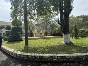 Bức ảnh chụp một khung cảnh ngoài trời trong một công viên hoặc khu tưởng niệm vào một ngày nắng. Ở phía trước là một bờ kè thấp bằng đá uốn cong, bao quanh một bãi cỏ xanh. Có nhiều cây xanh, trong đó có một cây lớn với phần gốc được sơn vôi trắng. Ở giữa bãi cỏ là một đài tưởng niệm bằng bê tông màu vàng nhạt có cấu trúc hình lục giác. Bên cạnh đó là một chiếc máy ủi cũ màu vàng được trưng bày. Phía sau là một tòa nhà màu vàng và nhiều cây cối khác. Bầu trời trong xanh và có mây.。（日本語訳）。写真は、晴れた日の公園または記念公園の屋外の風景を撮影したものです。手前には、緑の芝生を囲む、湾曲した低い石の縁石があります。多くの緑の木々があり、その中には根元が白く塗られた大きな木が一本あります。芝生の中央には、六角形の構造をした薄黄色のコンクリート製の記念碑があります。その隣には、古い黄色のブルドーザーが展示されています。後方には、黄色い建物と他の多くの木々があります。空は青く澄んでおり、雲があります。