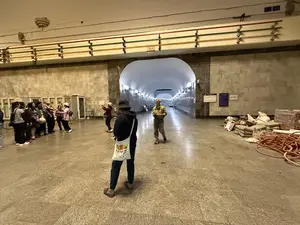 Bên trong một nhà ga tàu điện ngầm rộng lớn với kiến trúc trần vòm và tường ốp đá. Một nhóm người, có vẻ là khách du lịch, đang tụ tập ở phía bên trái, một số đang chụp ảnh. Ở trung tâm, một người mặc áo hoodie đen và đội mũ tai bèo đang đứng quay lưng lại. Một người đàn ông lớn tuổi mặc áo sơ mi kẻ sọc đang đi về phía trước. Lối đi dẫn vào một đường hầm sâu hơn được chiếu sáng bởi các ngọn đèn treo tường. Phía bên phải của nhà ga có vẻ đang được thi công, với các vật liệu xây dựng như bao tải và một cuộn ống màu cam nằm trên sàn. Trên tường phía trên có các đường ống được sơn màu đỏ, xanh lá và vàng.。（日本語訳）。アーチ型の天井と石張りの壁を持つ広大な地下鉄の駅構内。左側には、観光客と思われる一団が集まっており、何人かは写真を撮っている。中央には、黒いパーカーを着てバケットハットをかぶった人が背を向けて立っている。ストライプのシャツを着た年配の男性が前方へ歩いている。通路は、壁掛けのランプに照らされた、より深いトンネルへと続いている。上方の壁には、赤、緑、黄色に塗られたパイプがある。