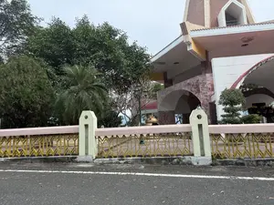 Bức ảnh chụp một con đường nhựa ở phía trước một công trình kiến trúc. Ngăn cách giữa đường và công trình là một hàng rào với các trụ màu xanh nhạt và lan can sắt sơn vàng có hoa văn độc đáo. Bên trong hàng rào là một khoảng sân và một tòa nhà có kiến trúc hiện đại, kết hợp giữa tường ốp đá, mái vòm và mái ngói. Xung quanh có nhiều cây xanh tươi tốt, bao gồm cả cây lớn và cây cọ. Bầu trời trong ảnh có màu xám, u ám. Phía sau hàng rào có thể thấy một chiếc lư hương lớn bằng đồng và một cây đèn đường kiểu cổ.。（日本語訳）。写真は、ある建築物の前にあるアスファルトの道を写しています。道と建築物を隔てているのは、水色の柱とユニークな模様がある黄色に塗られた鉄の手すりを持つ一列の柵です。柵の内側には中庭と、石張りの壁、アーチ型の屋根、瓦屋根を組み合わせた現代的な建築様式の建物があります。周りには、大きな木やヤシの木など、青々と茂った多くの木々があります。写真の中の空は灰色で、曇っています。柵の後ろには、大きな銅製の香炉とアンティーク調の街灯が見えます。