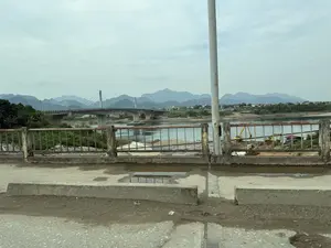 Bức ảnh chụp một phong cảnh miền núi với một con sông rộng. Ở phía xa, một cây cầu dây văng hiện đại bắc qua sông. Phía sau cây cầu là những dãy núi trập trùng và các ngôi nhà dân. Dọc bờ sông có một công trường xây dựng với máy xúc màu vàng. Ở tiền cảnh là lan can bằng bê tông và kim loại đã cũ, gỉ sét, và mặt đường bê tông có vũng nước. Bầu trời u ám và có nhiều mây.。（日本語訳）。写真は、広い川のある山岳風景を写しています。遠くには、近代的な斜張橋が川に架かっています。橋の後ろには、連なる山々と民家があります。川岸に沿って、黄色いショベルカーのある建設現場があります。手前には、古びて錆びたコンクリートと金属製の手すり、そして水たまりのあるコンクリートの路面があります。空はどんよりとしており、雲が多いです。