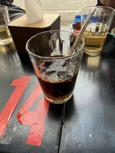 Trên một chiếc bàn màu đen, có một ly cà phê sữa chưa được khuấy, với lớp sữa đặc ở dưới đáy và cà phê đen ở trên. Trong ly có một chiếc thìa. Mặt bàn được dán số 14 bằng băng dính màu đỏ đã hơi bong tróc. Ở phía sau là một ly trà đá và một hộp khăn giấy.。（日本語訳）。黒いテーブルの上に、まだかき混ぜられていないミルクコーヒーが一杯あります。底にはコンデンスミルクの層があり、上にはブラックコーヒーがあります。グラスの中にはスプーンが一本入っています。テーブルの表面には、少し剥がれかけた赤い粘着テープで14という数字が貼られています。その後ろには、アイスティーが一杯とティッシュペーパーの箱が一つあります。