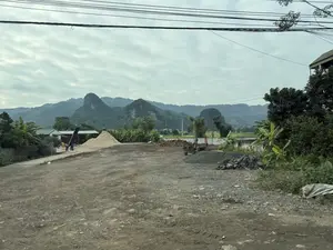 Bức ảnh chụp một khung cảnh ở vùng nông thôn miền núi. Tiền cảnh là một khoảng sân đất rộng, lổn nhổn sỏi đá, có một đống cát lớn màu trắng và một đống đá dăm nhỏ hơn. Có một vài máy móc công nghiệp và xe công nông. Ở phía sau, có những ngôi nhà và nhà xưởng với mái tôn xanh, bên cạnh là những lùm cây chuối và cây cối khác. Xa xa là những dãy núi đá vôi hùng vĩ, cây cối bao phủ xanh mướt. Bầu trời u ám, nhiều mây, và có nhiều đường dây điện chằng chịt ở phía trên.。（日本語訳）。写真は山岳地帯の田舎の風景を写したものです。前景は、砂利ででこぼこした広い土の庭で、大きな白い砂の山と、それより小さな砕石の山があります。いくつかの産業機械と耕運機があります。後方には、緑のトタン屋根の家や作業場があり、その隣にはバナナの木やその他の木々の茂みがあります。遠くには、青々とした木々に覆われた雄大な石灰岩の山々があります。空はどんよりと曇っており、上空には多くの電線が複雑に張り巡らされています。