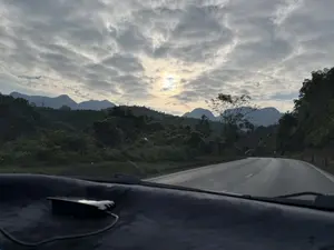 Bức ảnh được chụp từ bên trong một chiếc ô tô, nhìn ra con đường nhựa uốn lượn qua vùng đồi núi. Bầu trời đầy mây, mặt trời đang cố gắng chiếu những tia nắng xuyên qua. Phía xa là những dãy núi trập trùng. Hai bên đường là cây cối và thảm thực vật xanh tươi. Phía dưới bức ảnh là một phần của bảng điều khiển xe hơi.。（日本語訳）。空は雲に覆われ、太陽が雲間から光を差し込もうとしている。遠くには、幾重にも連なる山々がある。道の両側には木々と青々とした草木がある。