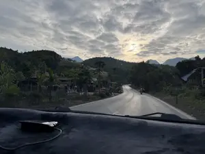 Bức ảnh được chụp từ bên trong một chiếc ô tô, nhìn ra một con đường nhựa uốn lượn ở vùng nông thôn miền núi. Bầu trời nhiều mây, mặt trời đang cố gắng chiếu xuyên qua những đám mây. Hai bên đường là những ngôi nhà, cây cối và những ngọn đồi xanh. Có một vài người đang đi xe máy trên đường. Bên trái có một ngôi nhà với quần áo đang phơi trên dây. Phía trước là một phần táp lô của xe ô tô.。（日本語訳）。空は雲が多く、太陽が雲を突き抜けようとしている。道の両側には家々、木々、そして緑の丘がある。道にはバイクに乗っている人が何人かいる。左側には、ロープに服が干されている家がある。