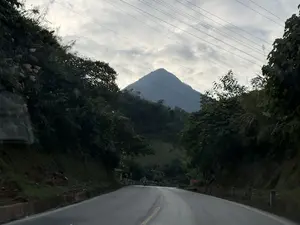 Bức ảnh chụp một con đường nhựa uốn lượn qua vùng núi. Hai bên đường là sườn đồi dốc đứng, được bao phủ bởi cây cối xanh tươi. Ở phía xa, một ngọn núi có đỉnh nhọn hình kim tự tháp hiện ra sừng sững dưới bầu trời u ám, đầy mây. Những đường dây điện chạy ngang qua khung cảnh. Phía trước trên con đường, có thể thấy bóng dáng một người đang lái xe máy.。（日本語訳）。山岳地帯を曲がりくねって通るアスファルトの道を写した写真。道の両側は、青々とした木々に覆われた急な丘の斜面です。遠くには、ピラミッド型の尖った頂を持つ山が、雲の多い曇り空の下にそびえ立っています。電線が風景を横切っています。道の先には、バイクを運転している人の姿が見えます。