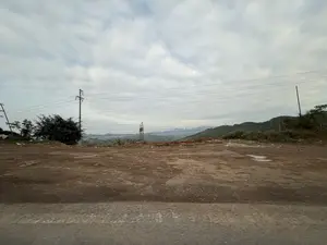 Bức ảnh chụp một khung cảnh vùng nông thôn vào một ngày nhiều mây. Bầu trời u ám, bao phủ bởi những đám mây trắng và xám. Ở phía trước là một khoảng đất trống rộng lớn, bề mặt là đất nâu, không bằng phẳng và có vài vũng nước nhỏ. Nhiều cột điện và dây điện chằng chịt chạy ngang qua khung cảnh. Ở phía xa là những dãy núi trập trùng được cây cối bao phủ, mờ ảo trong sương.。（日本語訳）。写真は曇りの日の田舎の風景を写しています。空はどんよりとしており、白と灰色の雲に覆われています。手前には広大な空き地があり、その表面は茶色い土で、平らではなく、いくつかの小さな水たまりがあります。多くの電柱と電線が風景を横切って複雑に走っています。遠くには木々に覆われた山々が連なり、霧の中にぼんやりと見えています。