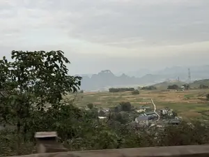 Phong cảnh một vùng quê miền núi nhìn từ trên cao. Bầu trời u ám và đầy mây. Phía xa, những dãy núi đá vôi trùng điệp ẩn hiện trong sương mù. Bên dưới là một thung lũng rộng lớn với những cánh đồng lúa, một số đã được thu hoạch có màu vàng nâu, xen kẽ với những mảng ruộng xanh. Một ngôi làng nhỏ nằm nép mình trong thung lũng, với những ngôi nhà có mái ngói màu xám và xanh lục. Một con đường đất nhỏ chạy quanh co qua làng và cánh đồng. Ở phía trước bên trái là một tán cây xanh rậm rạp, và ở dưới cùng là một phần của lan can đường.。（日本語訳）。高い場所から見た山岳地帯の田舎の風景。空はどんよりと曇っている。遠くには、幾重にも重なる石灰岩の山々が霧の中に姿を現したり隠したりしている。眼下には広大な谷が広がり、水田が広がっている。一部は収穫されて黄褐色になり、緑の田んぼと交互に並んでいる。谷間には小さな村がひっそりと佇み、灰色や緑色の瓦屋根の家々が建ち並んでいる。小さな未舗装の道が村や田んぼを曲がりくねって続いている。