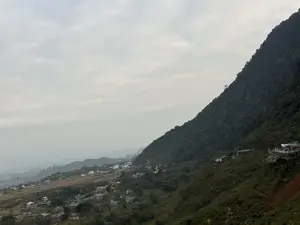 Bức ảnh chụp một khung cảnh vùng núi rộng lớn dưới bầu trời u ám, đầy mây. Một bên là sườn núi dốc, được bao phủ dày đặc bởi cây cối xanh um. Bên còn lại là một thung lũng, nơi có một ngôi làng nhỏ với những ngôi nhà nằm rải rác. Xa xa, có thể nhìn thấy những dãy núi mờ ảo và các cột điện cao thế. Trên sườn núi gần hơn, có một vài công trình kiến trúc trông giống như các điểm dừng chân hoặc quán cà phê ngắm cảnh dành cho khách du lịch.。（日本語訳）。写真は、曇り空の下に広がる広大な山岳風景を捉えている。片側には、青々とした木々で密に覆われた急な山の斜面がある。もう一方には谷があり、そこには家々が点在する小さな村がある。手前の山の斜面には、観光客向けの休憩所や展望カフェのような建築物がいくつかある。