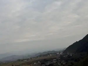 Bức ảnh chụp một khung cảnh vùng núi rộng lớn dưới bầu trời u ám, đầy mây. Phía dưới là một thung lũng với những ngôi nhà nằm rải rác trên sườn đồi, xen kẽ với cây cối và những mảnh đất trống. Một sườn núi lớn, rậm rạp cây xanh chiếm phần bên phải của bức ảnh. Nhìn ra xa, các dãy núi khác hiện ra mờ ảo trong lớp sương mù. Một vài cột điện cao thế cũng có thể được nhìn thấy trong khung cảnh.。（日本語訳）。写真は、曇り空の下、広大な山岳風景を写しています。下方には、丘の中腹に家々が点在し、木々や空き地が混在する谷があります。緑豊かな大きな山腹が写真の右側を占めています。遠くを見ると、他の山脈が霧の中にぼんやりと現れています。風景の中には、いくつかの高圧電線の鉄塔も見ることができます。