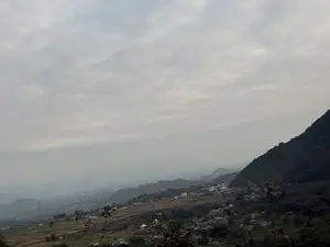 Bức ảnh chụp một khung cảnh vùng núi rộng lớn dưới bầu trời u ám, đầy mây. Phía bên phải là một sườn núi dốc đứng, cây cối rậm rạp. Nhìn xuống dưới là một thung lũng với một ngôi làng nhỏ, nơi có những ngôi nhà mái ngói nằm rải rác trên sườn đồi. Xung quanh làng là những mảnh đất canh tác và cây cối. Ở phía xa, những dãy núi khác hiện ra mờ ảo trong làn sương mù.。（日本語訳）。写真は、曇り空の下に広がる広大な山岳風景を写している。右側には、木々が鬱蒼と茂る急な山腹がある。見下ろすと、丘の中腹に瓦屋根の家々が点在する小さな村のある谷がある。村の周りには、耕作地と木々がある。遠くには、他の山々が霧の中にぼんやりと現れている。