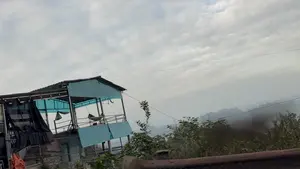 Bức ảnh chụp một ngôi nhà có kết cấu đơn giản, dựng trên sườn đồi. Ngôi nhà có khung kim loại, với phần mái và vách che bằng các tấm nhựa màu xanh lam. Tầng trên là một ban công mở, có một chiếc võng được mắc. Phía xa là khung cảnh núi non trập trùng chìm trong sương mù. Bầu trời u ám, đầy mây trắng xám.。（日本語訳）。写真には、丘の中腹に建てられた、シンプルな構造の家が写っている。家は金属製の骨組みで、屋根と壁は青いプラスチックの板で覆われている。上階は開かれたバルコニーで、ハンモックが1つ吊るされている。遠方には、霧に沈む幾重にも連なる山々の風景が広がっている。空はどんよりとしており、灰白色の雲で覆われている。