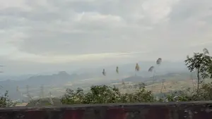 Phong cảnh núi non nhìn từ trên cao xuống một thung lũng. Bầu trời u ám, đầy mây. Phía xa là những dãy núi trập trùng chìm trong sương mù. Dưới thung lũng là những cánh đồng, làng mạc và các cột điện cao thế. Ở tiền cảnh là những bông cỏ lau đang bay trong gió bên cạnh một bờ tường bê tông.。（日本語訳）。上から谷を見下ろした山々の風景。空はどんよりとしており、雲に覆われている。遠くには、霧に沈む幾重にも連なる山々がある。谷の下には、田畑、村々、そして高圧電線の鉄塔がある。手前には、コンクリートの壁のそばで風に舞うススキの穂がある。