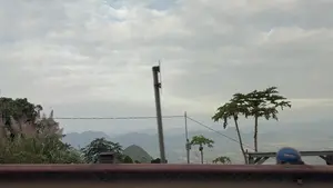 Bức ảnh chụp phong cảnh núi non hùng vĩ nhìn từ trên cao. Bầu trời u ám, đầy mây trắng xám. Phía xa là những dãy núi trùng điệp, lớp lớp nối tiếp nhau, mờ ảo trong sương. Bên dưới là một thung lũng rộng lớn. Ở gần hơn, có một vài cây đu đủ vươn cao. Một chiếc mũ bảo hiểm màu xanh có sọc đỏ và trắng được đặt trên một băng ghế gỗ ở bên phải. Một lan can màu nâu đỏ chạy ngang ở phía trước, và có một cột điện cùng những bụi cỏ lau ở bên trái.。（日本語訳）。高いところから見た雄大な山々の風景を写した写真。空はどんよりとしており、白と灰色の雲でいっぱいだ。遠くには、幾重にも連なる山々が霧の中にぼんやりと見え、次々と続いている。下には広大な谷が広がっている。手前には、高く伸びるパパイヤの木が数本ある。前方には赤茶色の手すりが横切っており、左側には電柱とススキの茂みがある。