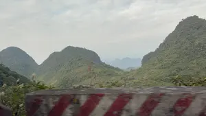 Bức ảnh chụp phong cảnh núi non hùng vĩ, nhìn từ một phương tiện đang di chuyển. Những dãy núi đá vôi trùng điệp, được bao phủ bởi cây cối xanh tươi, nối tiếp nhau đến tận chân trời. Bầu trời u ám, có nhiều mây và sương mù bao phủ các ngọn núi ở phía xa. Ở phía trước, một phần của hàng rào bê tông ven đường với các vạch sơn đỏ và trắng bị mờ nhòe.。（日本語訳）。移動中の乗り物から見た、雄大な山々の風景写真。青々とした木々に覆われた石灰岩の山々が幾重にも重なり、地平線の果てまで連なっている。空はどんよりとしており、遠くの山々は多くの雲と霧に覆われている。手前には、赤と白の線が塗られた道端のコンクリート柵の一部がぼやけている。