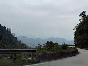 Khung cảnh một con đường đèo uốn lượn trong một ngày trời u ám. Phía xa là những dãy núi trập trùng ẩn hiện trong làn sương mờ. Dọc theo con đường là dải phân cách bằng bê tông và lan can bảo vệ. Sườn núi được bao phủ bởi cây cối rậm rạp. Vắt ngang bầu trời là những đường dây điện. Toàn cảnh mang vẻ tĩnh lặng và có phần hoang sơ của vùng núi.。（日本語訳）。曇りの日の、曲がりくねった峠道の風景。遠くには、薄い霧の中に幾重にも連なる山々が見え隠れしている。道に沿ってコンクリートの中央分離帯とガードレールがある。山の斜面は鬱蒼とした木々で覆われている。空を電線が横切っている。全景は、山岳地帯の静寂と、どこか手付かずの趣を帯びている。