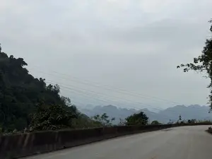 Phong cảnh một con đường đèo núi. Con đường nhựa uốn cong về phía bên phải, dọc theo đó là dải phân cách bằng bê tông. Bên trái là một ngọn đồi được bao phủ bởi cây cối rậm rạp. Phía xa, những dãy núi đá vôi trùng điệp hiện ra mờ ảo dưới bầu trời xám xịt, đầy mây. Vắt ngang qua bầu trời là những đường dây điện. Có một vài biển báo giao thông ở phía xa bên phải con đường.。（日本語訳）。山の峠道の風景。アスファルトの道は右側へカーブしており、それに沿ってコンクリート製の分離帯がある。左側は、鬱蒼とした木々に覆われた丘である。遠くには、雲が多くどんよりとした灰色の空の下、幾重にも重なる石灰岩の山々がぼんやりと姿を現している。空を電線が横切っている。道の右側の遠くに、いくつかの交通標識がある。