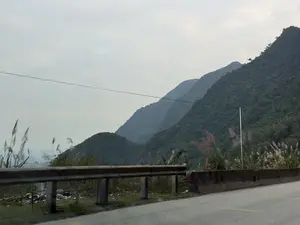 Bức ảnh chụp phong cảnh núi non từ một con đường. Ở phía trước là một đoạn đường có lan can bảo vệ bằng kim loại và dải phân cách bê tông. Ven đường mọc nhiều cỏ lau. Phía sau là những dãy núi trập trùng, cây cối xanh rậm rạp bao phủ. Những ngọn núi ở xa chìm trong màn sương mờ ảo. Bầu trời màu xám, u ám, cho thấy một ngày nhiều mây.。（日本語訳）。ある道から撮影された山々の風景写真。手前には、金属製のガードレールとコンクリートの中央分離帯がある道路の一部がある。道端にはススキがたくさん生えている。後ろには、青々とした木々で覆われた、幾重にも連なる山々がある。遠くの山々は幻想的な霧の中に沈んでいる。空は灰色でどんよりとしており、曇りの日であることを示している。