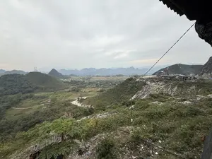 Bức ảnh chụp một khung cảnh thiên nhiên vùng núi rộng lớn, nhìn từ trên cao xuống dưới một bầu trời u ám, đầy mây. Phía trước là những ngọn đồi xanh trập trùng được bao phủ bởi cây cối. Xa xa, những dãy núi đá vôi nối tiếp nhau mờ ảo trong sương. Dưới thung lũng là những cánh đồng, vài ngôi nhà nằm rải rác và một con đường đất. Nổi bật trên sườn đồi bên phải là một khu nhà đang được xây dựng dở dang. Một cột điện cao thế cũng có thể được nhìn thấy trên một đỉnh đồi. Ở tiền cảnh, có cây cối, bao gồm cả cây đu đủ, nhưng mặt đất lại có nhiều rác thải vương vãi.。（日本語訳）。写真は、曇り空の下、広大な山岳地帯の自然風景を上から見下ろして撮影したものである。手前には、木々に覆われた緑の丘が幾重にも連なっている。遠くには、石灰岩の山々が霧の中にぼんやりと連なっている。谷の下には、田畑、点在する数軒の家、そして未舗装の道がある。右側の丘の中腹には、建設途中の建物群が目立っている。丘の頂上には高圧電線の鉄塔も見られる。前景には、パパイヤの木を含む木々があるが、地面には多くのゴミが散乱している。