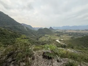 Bức ảnh chụp một khung cảnh thiên nhiên rộng lớn ở vùng núi, nhìn từ trên cao xuống. Phía trước là một sườn đồi dốc với đất đá, có vài cây đu đủ và một túp lều nhỏ tạm bợ, xung quanh có rác. Nhìn ra xa là một thung lũng xanh tươi với những ngọn đồi nhấp nhô, cây cối rậm rạp. Xen kẽ trong thung lũng là vài ngôi nhà, những mảnh ruộng và một con đường đất. Một dòng sông cạn với lòng sông màu sáng chảy qua khu vực. Phía chân trời là những dãy núi trập trùng mờ ảo trong sương. Bầu trời u ám, đầy mây.。（日本語訳）。写真は、山岳地帯の広大な自然風景を上から見下ろしたものです。手前には土石のある急な丘の斜面があり、パパイヤの木が数本と小さな仮設小屋が一つあり、周りにはゴミがあります。遠くには、起伏のある丘と鬱蒼とした木々がある緑豊かな谷が見えます。谷間には、数軒の家、田畑、そして未舗装の道が点在しています。明るい色の川床を持つ、水量の少ない川がこの地域を流れています。空はどんよりとしており、雲でいっぱいです。