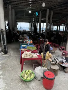 Bức ảnh chụp một quán ăn ven đường, có thể là ở một khu chợ vùng cao. Một người phụ nữ đang ngồi trên chiếc ghế nhựa nhỏ bên cạnh bếp than đang cháy. Nhiều bắp ngô nướng được xiên vào que và cắm gần bếp. Trên những chiếc bàn nhựa, các món ăn được bày bán, bao gồm rất nhiều ống cơm lam, một đĩa xúc xích và nem nướng đặt trên lá chuối. Ngoài ra còn có một rổ ngô tươi, một bát trứng và nhiều hũ đồ ngâm. Khung cảnh xung quanh khá đơn sơ với nền xi măng, các cột trụ bê tông và mái che. Phía xa có thể thấy dãy núi mờ ảo.。（日本語訳）。写真は道端の食堂を写したもので、おそらく山岳地帯の市場にあるものだろう。一人の女性が、燃えている炭火コンロの隣にある小さなプラスチックの椅子に座っている。多くの焼きトウモロコシが串に刺され、コンロの近くに立てられている。プラスチックのテーブルの上には、たくさんのコムラム（竹筒ご飯）、バナナの葉の上に置かれたソーセージとネムヌォン（焼きつくね）の皿など、料理が並べられて売られている。その他にも、生のトウモロコシが入ったかご、卵の入ったボウル、そして多くの漬物の瓶がある。周囲の風景は、セメントの床、コンクリートの柱、そして屋根で、かなり質素である。
