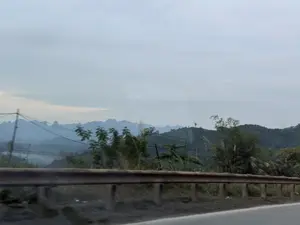 Bức ảnh chụp phong cảnh miền núi nhìn từ một con đường. Phía trước là lan can bảo vệ bằng kim loại chạy dọc theo mép đường. Bên kia lan can là cây cối và thảm thực vật. Ở phía xa, những dãy núi trập trùng với các đỉnh nhọn hiện lên mờ ảo trong sương. Bầu trời có màu xám nhạt, u ám, tạo nên một không gian tĩnh lặng.。（日本語訳）。ある道から見た山岳風景の写真。手前には、道路の縁に沿って走る金属製のガードレールがある。ガードレールの向こう側には木々と植生がある。遠くには、鋭い頂を持つ幾重にも重なる山脈が霧の中にぼんやりと現れている。空は薄い灰色で曇っており、静かな空間を作り出している。