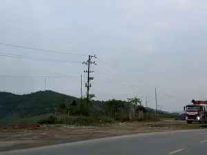 Bức ảnh chụp một khung cảnh ven đường ở một vùng núi. Bầu trời u ám, màu xám. Ở phía trước là một con đường nhựa. Bên lề đường, một chiếc xe tải lớn có cần cẩu màu đỏ và trắng đang đỗ. Có một cột điện lớn ở giữa với nhiều dây điện chằng chịt. Phía sau là những ngọn đồi xanh phủ đầy cây cối.。（日本語訳）。写真は山間部の道端の風景を撮影したものです。空は曇っており、灰色です。手前にはアスファルトの道路があります。道端には、赤と白のクレーンが付いた大きなトラックが停まっています。中央には大きな電柱があり、たくさんの電線が張り巡らされています。後ろには木々で覆われた緑の丘があります。