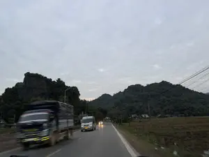 Khung cảnh trên một con đường ở vùng nông thôn miền núi vào lúc chạng vạng. Một chiếc xe tải màu xanh lam đang di chuyển nhanh ở bên trái, hình ảnh bị nhòe do chuyển động. Phía trước là một chiếc xe khách nhỏ màu bạc và một chiếc ô tô khác đang bật đèn pha ở phía xa. Hai bên đường là những dãy núi trập trùng được bao phủ bởi cây cối xanh tươi. Bên phải đường là một cánh đồng đã gặt, xa xa có vài ngôi nhà. Bầu trời u ám, đầy mây xám.。（日本語訳）。山間部の田舎道、夕暮れ時の光景。左側では青いトラックが速く移動しており、その動きで画像がぶれている。道路の両側には、緑の木々に覆われた起伏のある山々が連なっている。空は薄暗く、灰色の雲で覆われている。