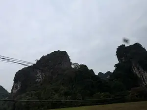 Phong cảnh núi đá vôi hùng vĩ dưới bầu trời u ám, đầy mây. Các ngọn núi được bao phủ bởi cây cối xanh rậm. Phía trước có một vài đường dây điện vắt ngang qua khung hình.。（日本語訳）。曇り空の下、雄大な石灰岩の山の風景。山々は青々と茂った木々で覆われている。手前には、数本の電線が画面を横切っている。