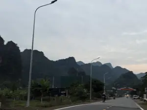 Phong cảnh một con đường ở khu vực miền núi vào lúc trời chập choạng tối. Bầu trời u ám, có nhiều mây. Phía xa là những dãy núi đá vôi trùng điệp. Dọc theo con đường là các cột đèn đường cao. Có một vài người đi xe máy và xe ô tô đang di chuyển trên đường. Cây cối mọc hai bên đường.。（日本語訳）。夕暮れ時の山間部にある一本道の風景。空は薄暗く、雲が多い。遠くには、幾重にも連なる石灰岩の山々がある。道沿いには高い街灯が並んでいる。数台のバイクと車が道路を移動している。道路の両脇には木々が生えている。