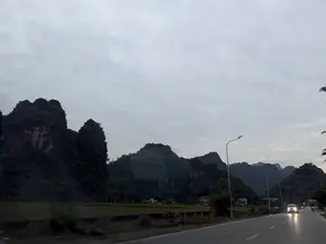 Phong cảnh ven đường vào buổi chiều tà với bầu trời xám xịt, nhiều mây. Nổi bật là những dãy núi đá vôi hùng vĩ, sẫm màu, được cây cối bao phủ. Bên cạnh là một con đường nhựa có các cột đèn đường. Một chiếc ô tô đang di chuyển trên đường và bật đèn pha. Ở phía xa, có những cánh đồng và vài ngôi nhà nằm dưới chân núi.。（日本語訳）。どんよりと雲の多い、夕暮れ時の道沿いの風景。際立っているのは、木々に覆われた、雄大で濃い色の石灰岩の山々だ。隣には街灯のあるアスファルトの道がある。一台の車がヘッドライトをつけて道を走っている。遠くには、山の麓に田畑といくつかの家がある。