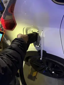 Một người đang cắm sạc cho một chiếc ô tô điện màu trắng. Người này mặc một chiếc áo khoác phao màu đen, tay đang giữ đầu sạc cắm vào cổng sạc của xe. Cảnh tượng diễn ra vào ban đêm hoặc trong một không gian tối, được chiếu sáng bởi một luồng ánh sáng vàng. Có thể nhìn thấy một phần bánh xe màu đen và đèn hậu màu đỏ của chiếc xe.。（日本語訳）。一人の人が白い電気自動車に充電している。この人は黒いダウンジャケットを着ており、充電プラグを手に持って車の充電ポートに差し込んでいる。光景は夜間または暗い空間で繰り広げられており、黄色い光に照らされている。