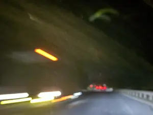 Bức ảnh mờ ảo, chụp vào ban đêm từ một phương tiện đang di chuyển, có thể là ô tô. Hình ảnh ghi lại những vệt sáng kéo dài trên một con đường tối, có thể là trong một đường hầm. Ở phía bên trái, các vệt sáng màu trắng và vàng của đèn pha tạo thành những đường dài. Ở phía xa, có thể thấy lờ mờ ánh đèn hậu màu đỏ của một chiếc xe khác. Bức ảnh tạo ra cảm giác mạnh mẽ về tốc độ và sự chuyển động trong không gian tối.。（日本語訳）。ぼやけた写真で、夜間に移動中の乗り物、おそらく自動車から撮影されたものである。この画像は、暗い道路、おそらくトンネル内で、長く伸びる光の筋を捉えている。左側では、ヘッドライトの白と黄色の光の筋が長い線を描いている。この写真は、暗い空間の中でのスピード感と動きを強く感じさせる。