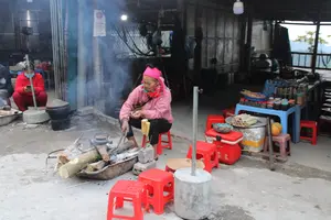 Trên tấm biển có chữ 'Cà phê' và 'VỆ SINH'. Bức ảnh chụp cảnh một quán ăn ven đường ở vùng cao. Một người phụ nữ mặc áo khoác hồng, đội khăn hồng, đang ngồi trên ghế nhựa đỏ và nướng đồ ăn bằng bếp củi. Khói bốc lên từ bếp. Gần đó có vài bắp ngô nướng. Ở phía sau, một người mặc đồ đỏ, đeo khẩu trang đang ngồi xem điện thoại. Một chiếc bàn bày bán các món ăn như thịt xiên, cơm lam và nhiều loại gia vị đựng trong hũ.。（日本語訳）。看板には「Cà phê（カフェ）」と「VỆ SINH（トイレ）」という文字がある。写真は、高原地帯の道端にある飲食店の光景を写している。ピンクの上着を着て、ピンクのスカーフを巻いた女性が、赤いプラスチックの椅子に座り、薪ストーブで食べ物を焼いている。ストーブから煙が立ち上っている。その近くには、焼いたトウモロコシが数本ある。後ろでは、赤い服を着てマスクをした人が座って携帯電話を見ている。テーブルには、串焼き肉、コムラム（竹筒ご飯）、そして瓶に入った多くの種類の調味料などの料理が並べられて売られている。