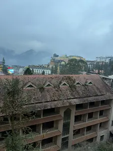 Chữ viết trong ảnh: SUNW... FANSIP..., SAPACENTRE, Hotel Sapa Square. Đây là quang cảnh thị trấn Sa Pa từ trên cao vào một ngày trời nhiều mây và sương mù. Ở tiền cảnh là mái ngói màu nâu của một tòa nhà đã phủ rêu. Phía xa là các công trình kiến trúc đa dạng của thị trấn, và những dãy núi chìm trong sương.。（日本語訳）。写真の中の文字：SUNW... FANSIP..., SAPACENTRE, Hotel Sapa Square。これは、雲と霧が多い日に上から見たサパの町の光景です。前景には、苔で覆われた建物の茶色い瓦屋根があります。遠くには、町の多様な建築物と、霧に沈む山々があります。