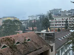 Quang cảnh một thị trấn trên sườn đồi chìm trong sương mù. Nhiều tòa nhà, khách sạn với kiến trúc đa dạng được xây dựng san sát nhau. Ở phía trước là mái nhà lợp ngói đỏ đã cũ và mọc rêu, có những cửa sổ nhỏ trên mái. Xen kẽ giữa các công trình là những hàng cây xanh. Bầu trời xám đặc, tạo nên một khung cảnh yên tĩnh của vùng cao.。（日本語訳）。霧に沈む丘の中腹にある町の光景。多様な建築様式の多くの建物やホテルが、密集して建てられている。手前には、古びて苔が生えた赤瓦の屋根があり、屋根には小さな窓がある。建造物の間には緑の木々が点在している。空はどんよりとした灰色で、高原の静かな風景を作り出している。