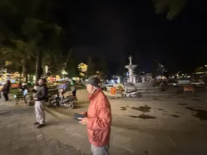 Bức ảnh chụp cảnh một quảng trường vào ban đêm, có vẻ khá đông người. Ở phía trước, một người đàn ông mặc áo khoác màu cam đỏ và đội mũ lưỡi trai đang đứng. Xung quanh có nhiều người khác, một vài chiếc xe máy đang đậu. Ở trung tâm là một đài phun nước lớn bằng đá màu trắng. Phía sau là cây cối và các tòa nhà sáng đèn dưới bầu trời đêm.。（日本語訳）。夜の広場の風景を撮った写真で、かなり混雑しているようです。前景には、赤みがかったオレンジ色のジャケットを着て野球帽をかぶった男性が立っています。周りには他の多くの人々がおり、数台のバイクが駐車しています。中央には、白い石でできた大きな噴水があります。後ろには木々や、夜空の下で明かりが灯る建物があります。