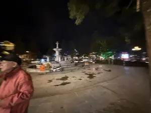 Bức ảnh chụp một quảng trường vào ban đêm, hình ảnh bị mờ do chuyển động. Ở trung tâm là một đài phun nước bằng đá. Sàn quảng trường lát đá và có nhiều vũng nước. Phía bên trái là một người đàn ông lớn tuổi mặc áo khoác màu đỏ và đội mũ đen. Gần đài phun nước có một chiếc xe máy đang đậu. Xung quanh là cây cối và các tòa nhà với nhiều ánh đèn đường và đèn trang trí bị nhòe trong đêm.。（日本語訳）。夜の広場を撮影した写真で、動きによって画像がぼやけている。中央には石造りの噴水がある。広場の床は石畳で、水たまりがたくさんある。左側には、赤い上着を着て黒い帽子をかぶった年配の男性がいる。噴水の近くにバイクが1台停まっている。周囲には木々や建物があり、多くの街灯や装飾の光が夜の中ににじんでいる。